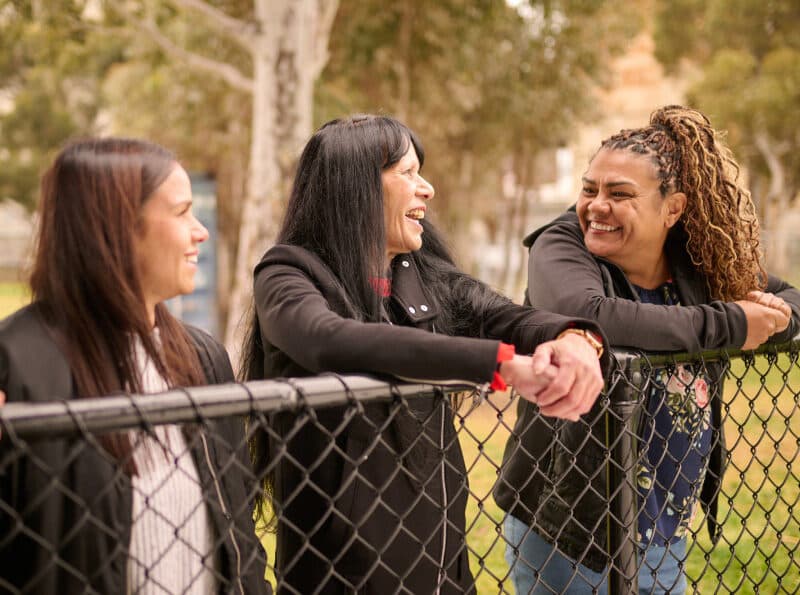 Three First Nations women chatting outdoors, leaning against a fence.