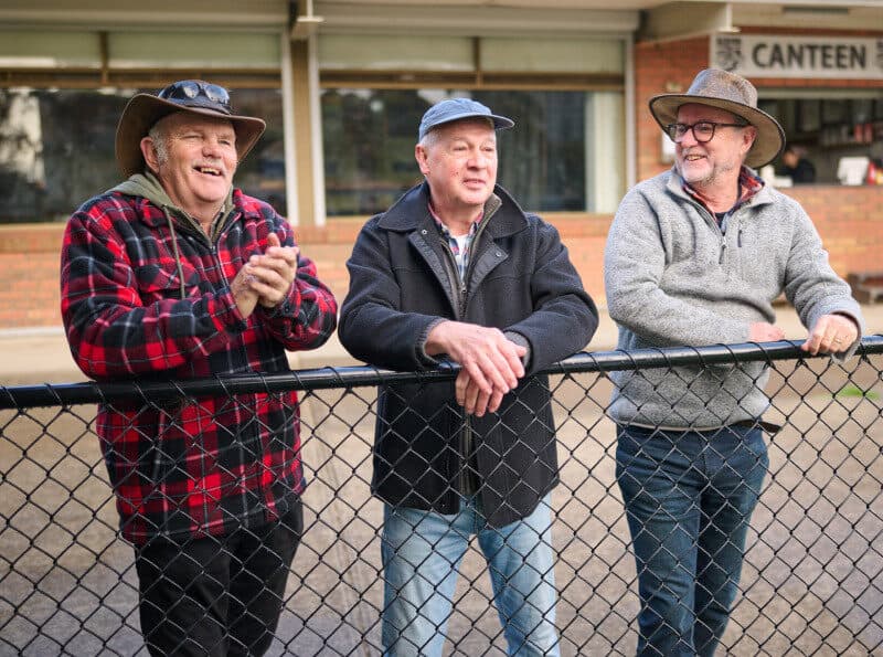 Three men chatting outdoors, leaning against a fence at a football match.