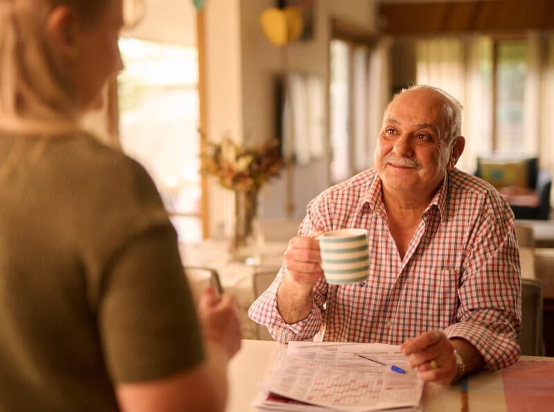 A man and woman chatting at the kitchen table. The man is holding a cup of coffee.